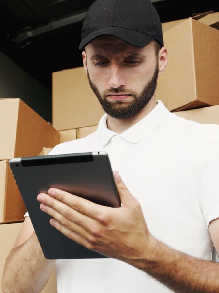 A service technician in a cap checking a job on a tablet beside stacked cartons