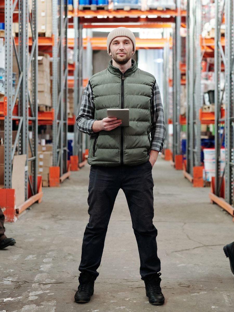 A warehouse lead holding a tablet in an aisle of tall pallet racks
